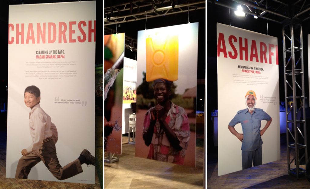 Tall exhibition panels with portraits of Chandresh, a woman carrying a jerry can, and Asharfi