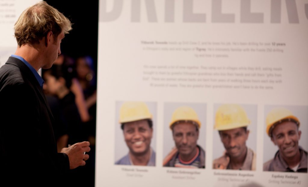 Close-up of an exhibition panel with portraits of four drillers in yellow hard hats, with a guest looking on